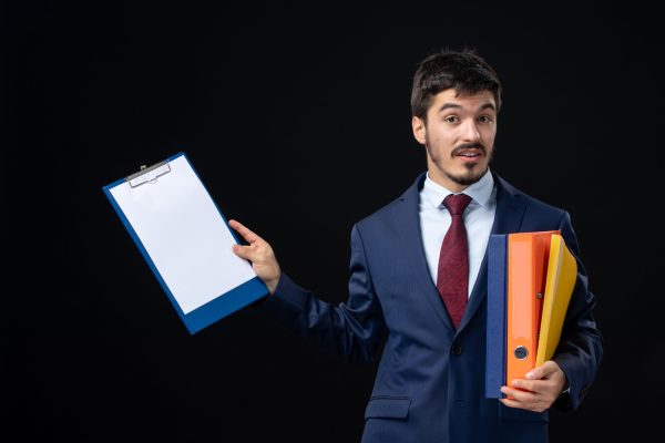 confused-male-suit-holding-several-documents-asking-something-isolated-dark-wall